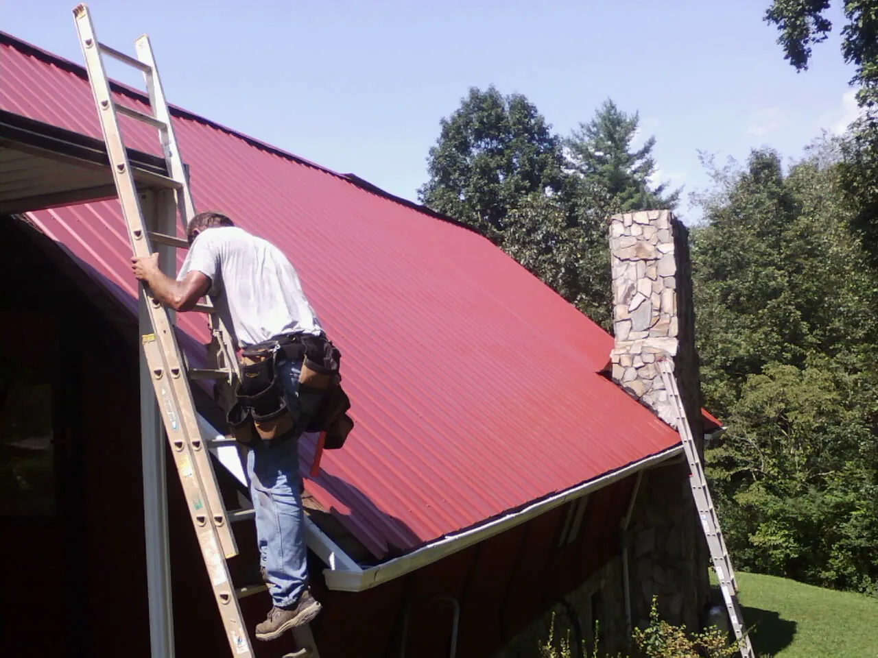 Roofer on a ladder beside a red metal roof and stone chimney — Soffit & Fascia Repair by Roofing Brothers in Taloga